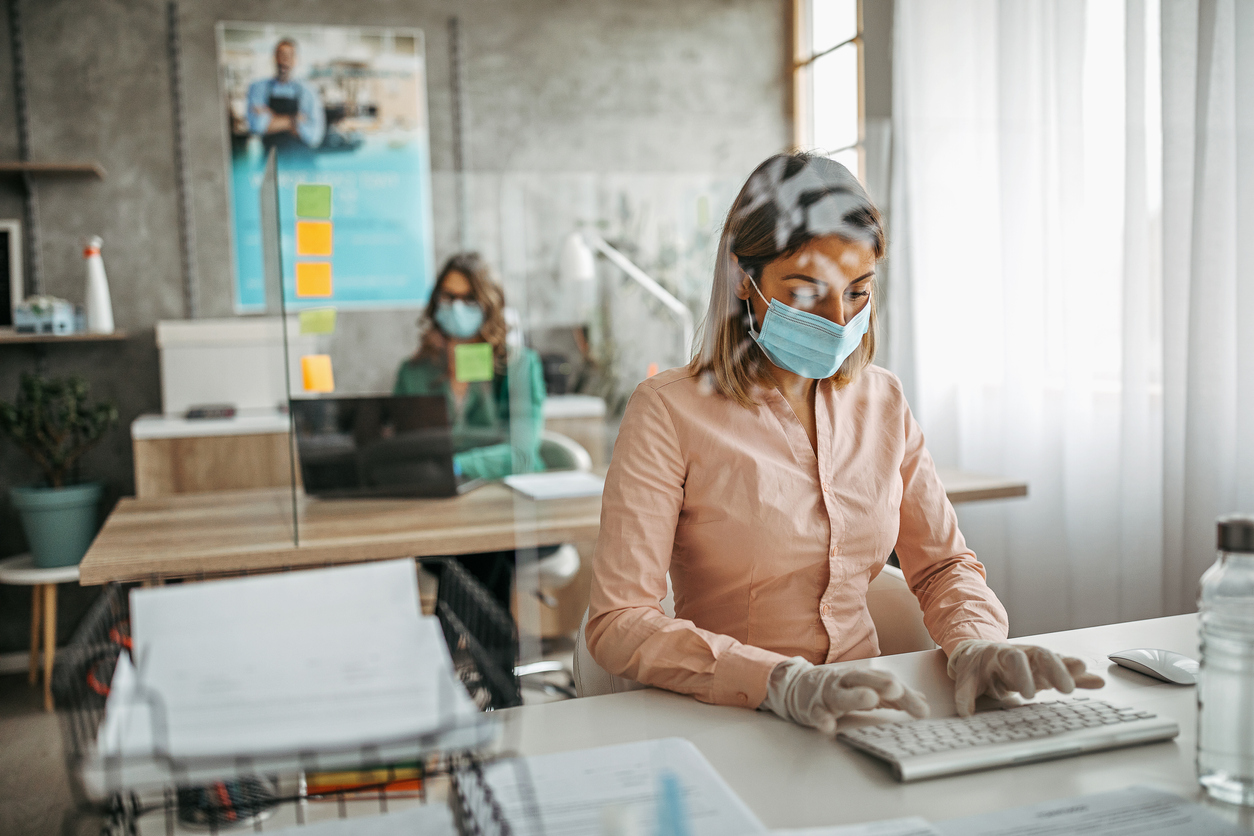 Two businesswomen working at office wearing protective gloves and face mask maintaining social distance. Office with acrylic glass partition on desk. Acrylic glass wall - protection against coughs and spitting, protection against viruses.